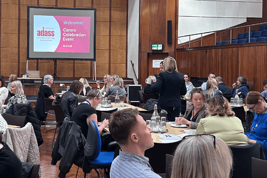 A large group of people gathered in a conference room, seated together around tables and standing chatting. A projector screen display at the front of the room reads: Welcome! Carers Celebration Event, 18/04/26.