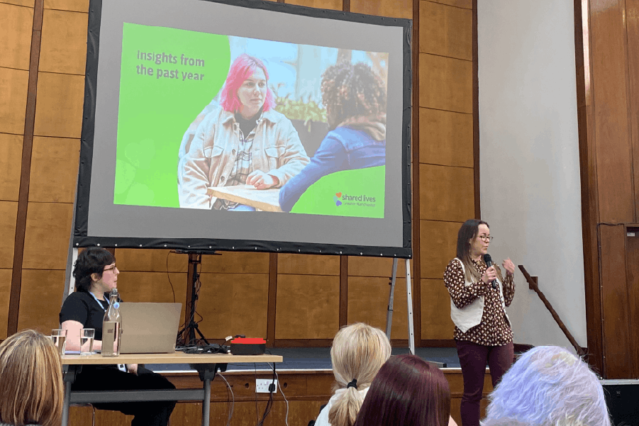 Jo stands in front of a projector screen, speaking animatedly into a microphone as seated people watch on. The screen displays an image of two people chatting at an outdoor table, with text that reads: Insights from the past year.