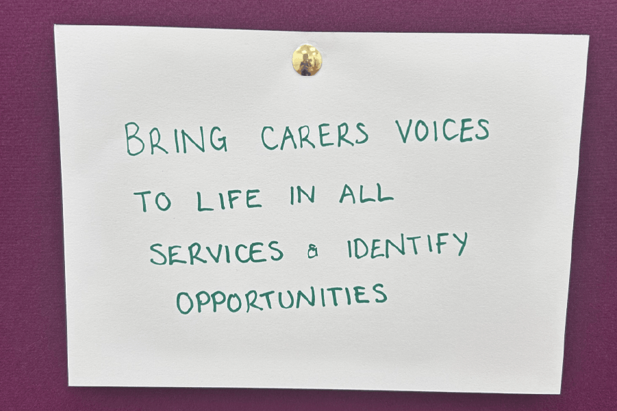 A white sheet of A4 paper pinned to a purple background with a brass tack. Handwriting in green pen reads: ‘Bring carers voices to life in all services and identify opportunities.’